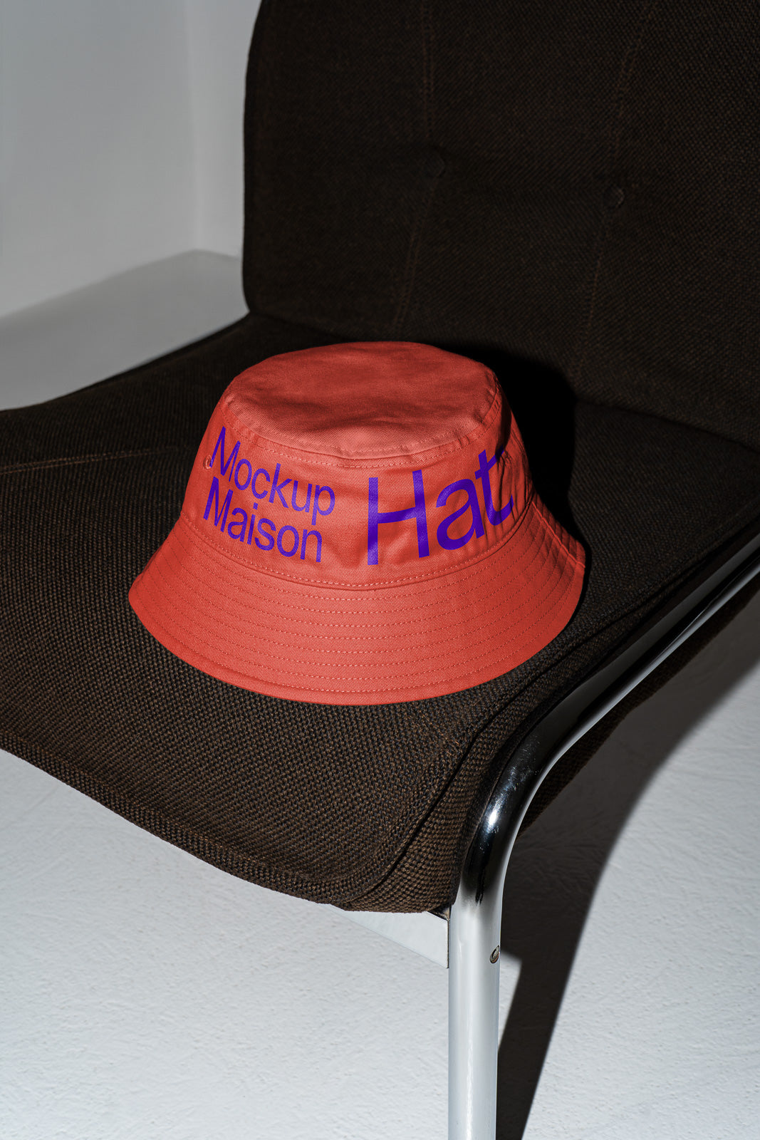 PSD mockup of an orange bucket hat with "Mockup Maison Hat" text in blue, placed on a dark textured chair with metal legs against a light floor.