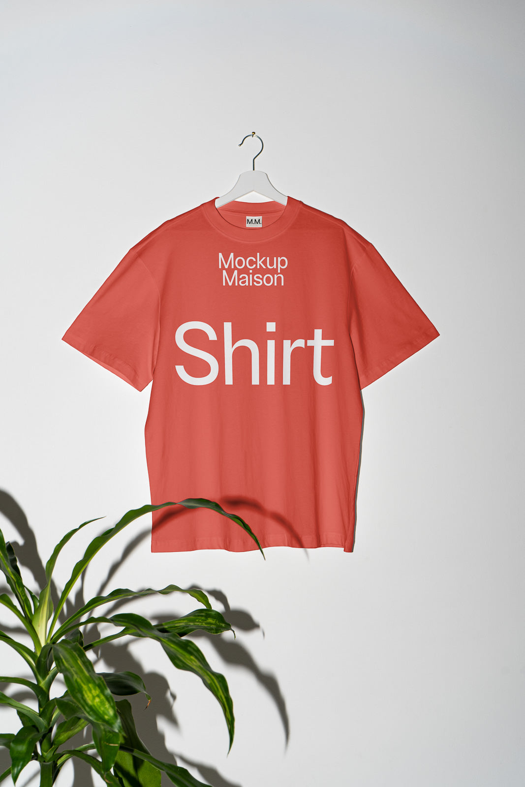 PSD mockup of a red T-shirt with "Mockup Maison Shirt" text hanging on a white hanger against a gray background with a green plant visible at the bottom left.