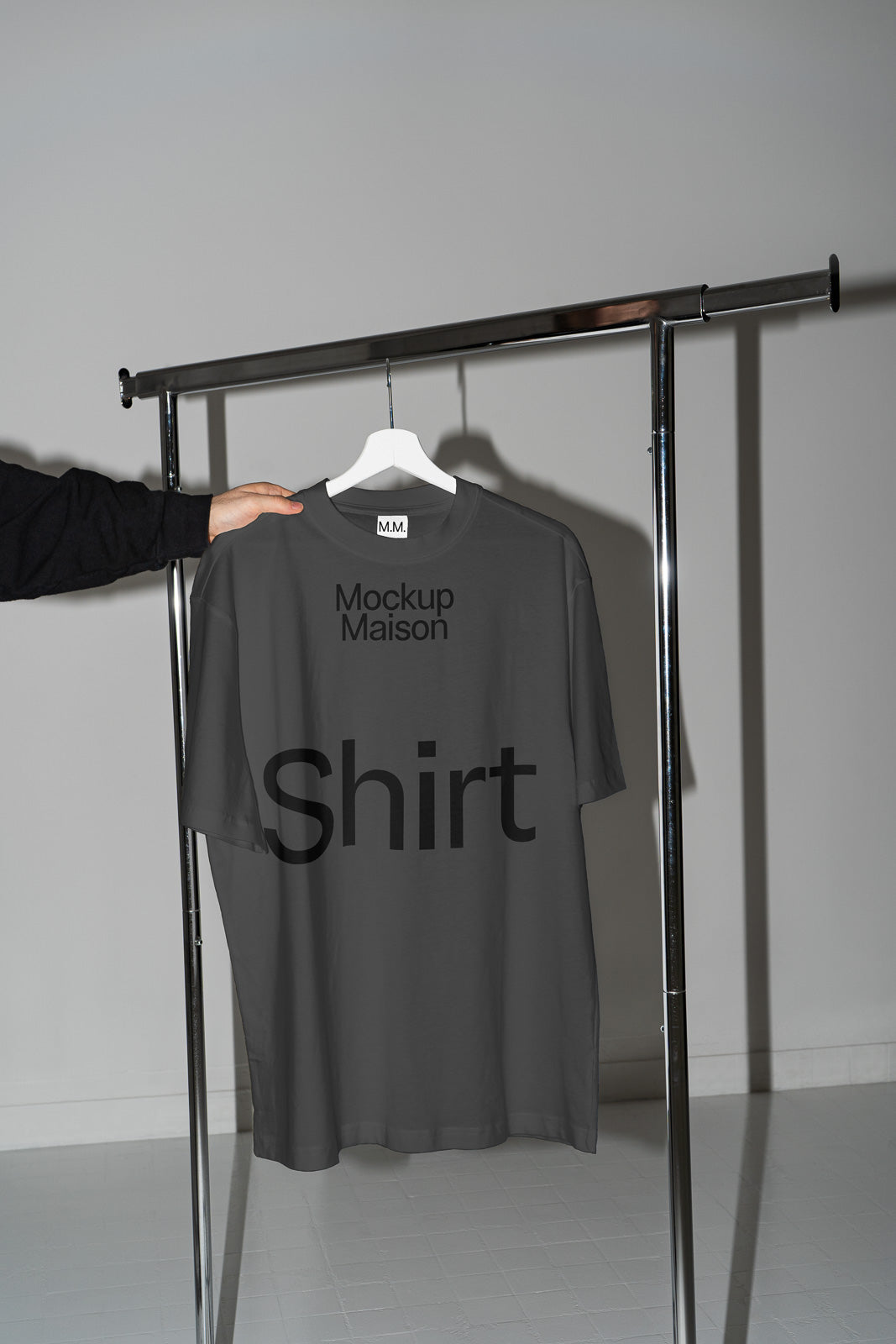 PSD mockup of a gray t-shirt labeled "Mockup Maison" on a hanger, held by a hand, displayed on a metal garment rack in a minimalistic setting.