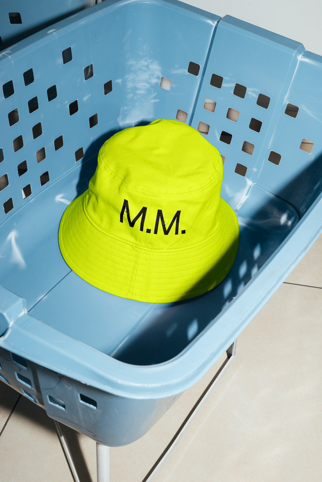 PSD mockup of a bright yellow bucket hat with "M.M." text, placed inside a blue perforated plastic basket on a tiled floor.