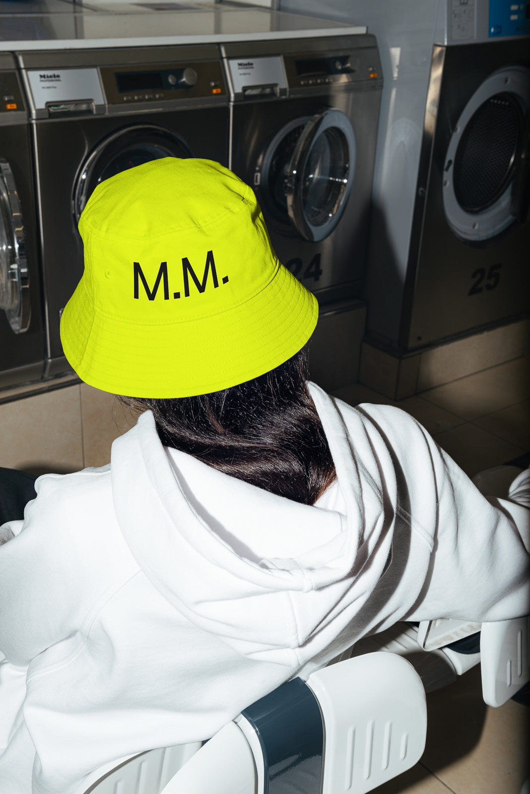 PSD mockup of a person with dark hair sitting in a laundromat, wearing a neon yellow bucket hat with the initials "M.M." and a white hoodie.