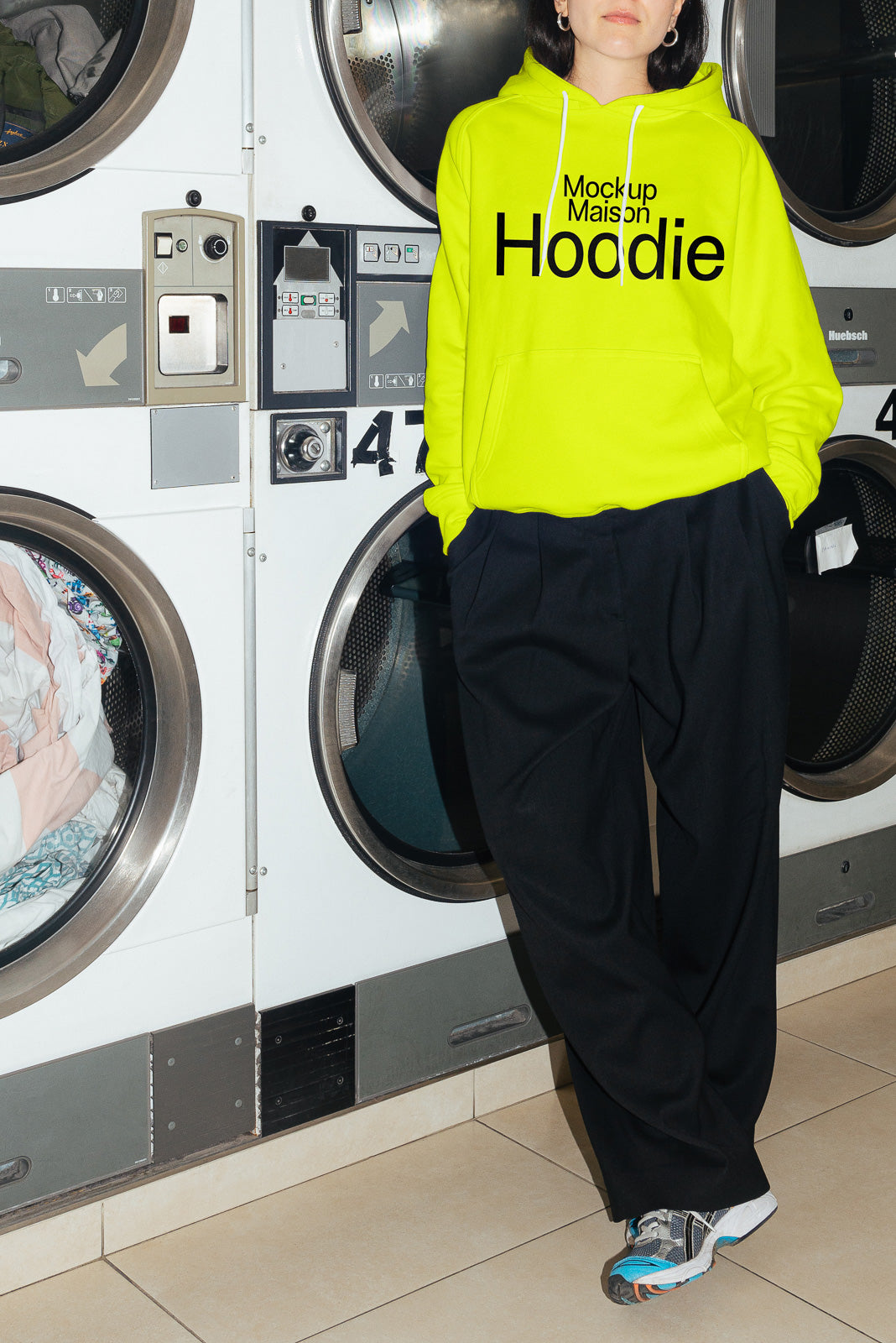 PSD mockup of a person wearing a neon green hoodie with text, standing in front of industrial washing machines.
