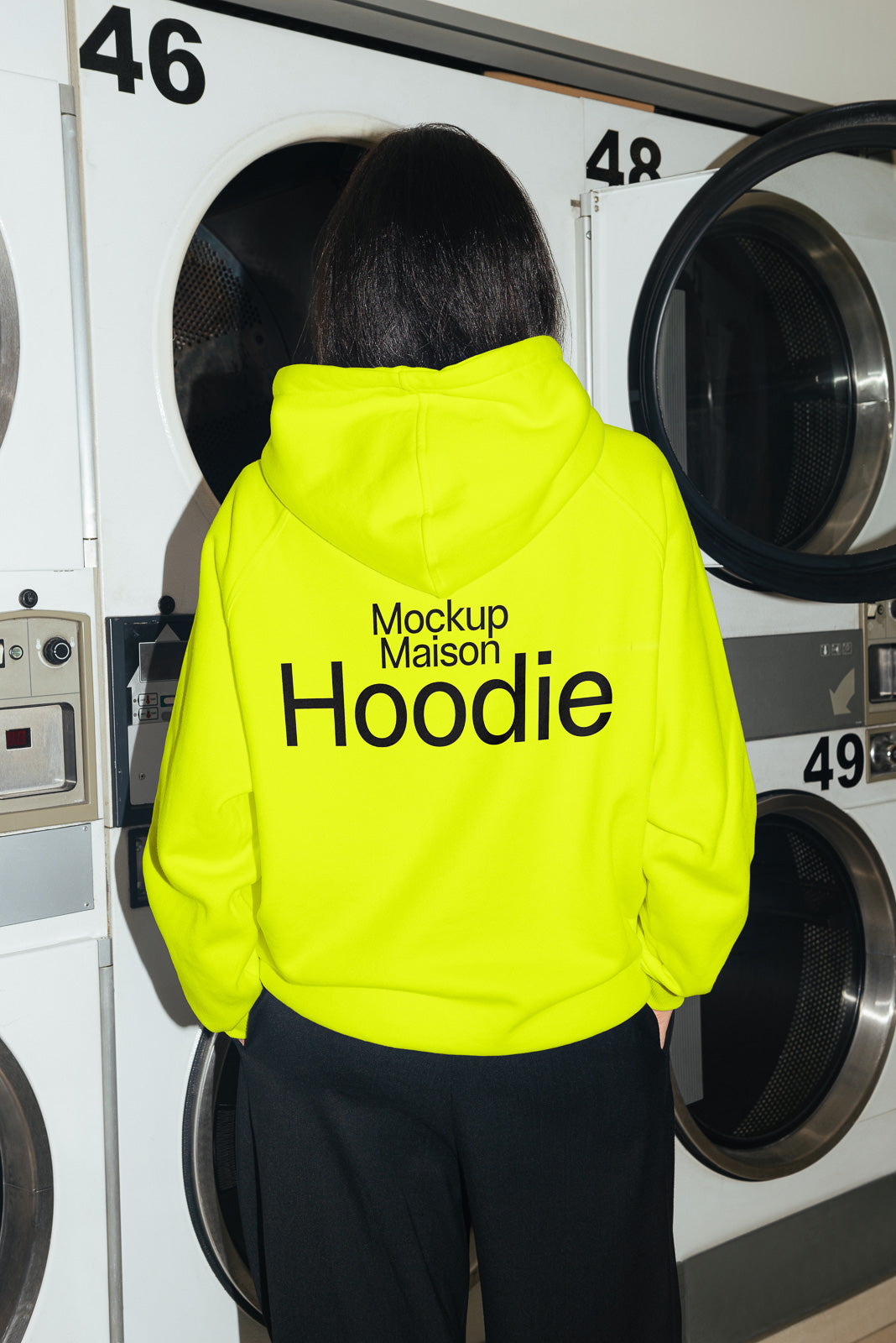 PSD mockup of a person standing with their back facing, wearing a bright yellow hoodie with text, in front of large industrial dryers marked with numbers.