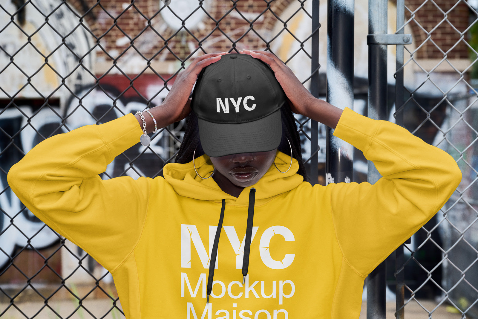 PSD mockup of a person in a yellow hoodie and black cap labeled "NYC," standing in front of a chain-link fence with graffiti.