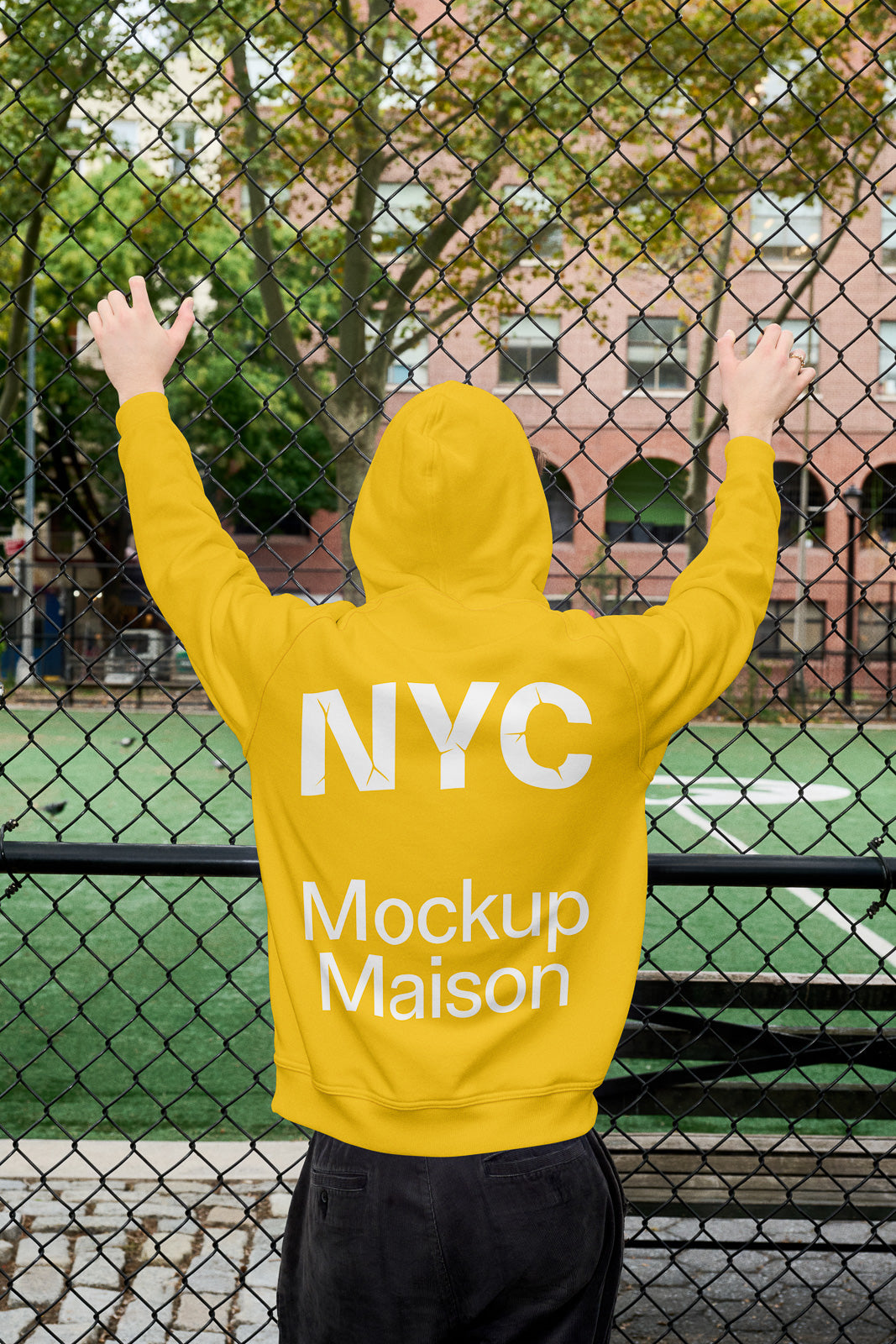 PSD mockup of a person wearing a yellow hoodie with "NYC Mockup Maison" text, standing by a chain-link fence with a green field beyond.