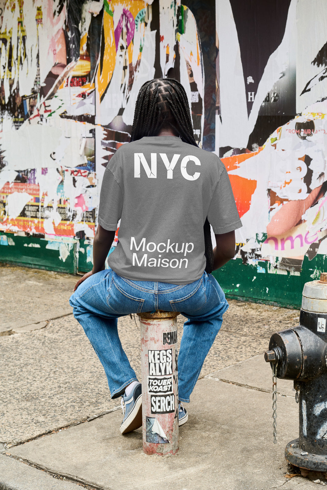 PSD mockup of a person wearing a gray T-shirt with "NYC Mockup Maison" printed on the back, sitting on a graffiti-covered post.