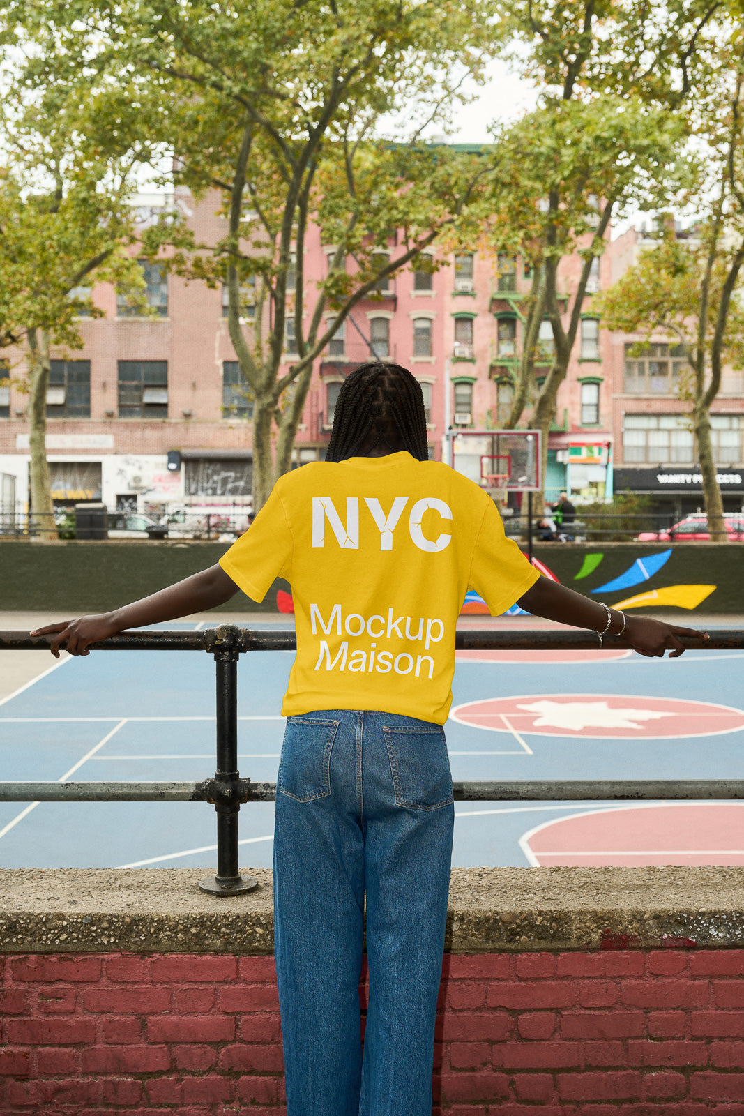 PSD mockup of a person wearing a bright yellow NYC Mockup Maison T-shirt, overlooking an urban basketball court with colorful murals.