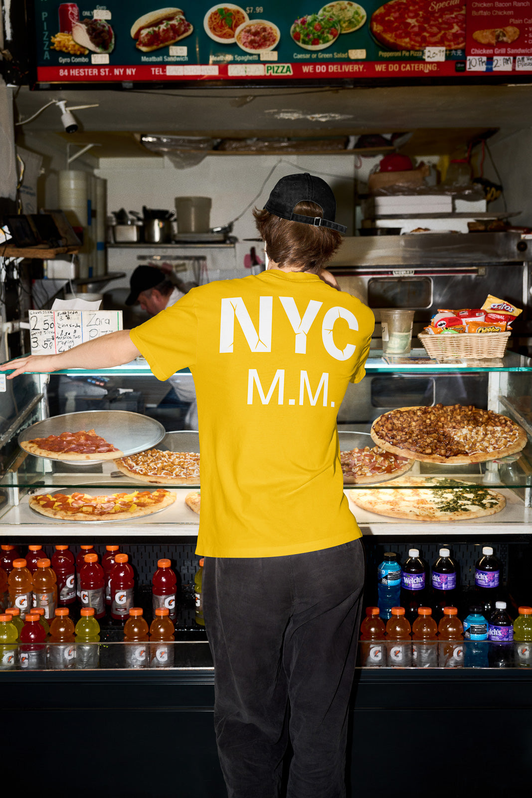 PSD mockup of a person wearing a yellow NYC shirt at a pizza counter with various pizzas on display and an overhead menu listing different dishes.