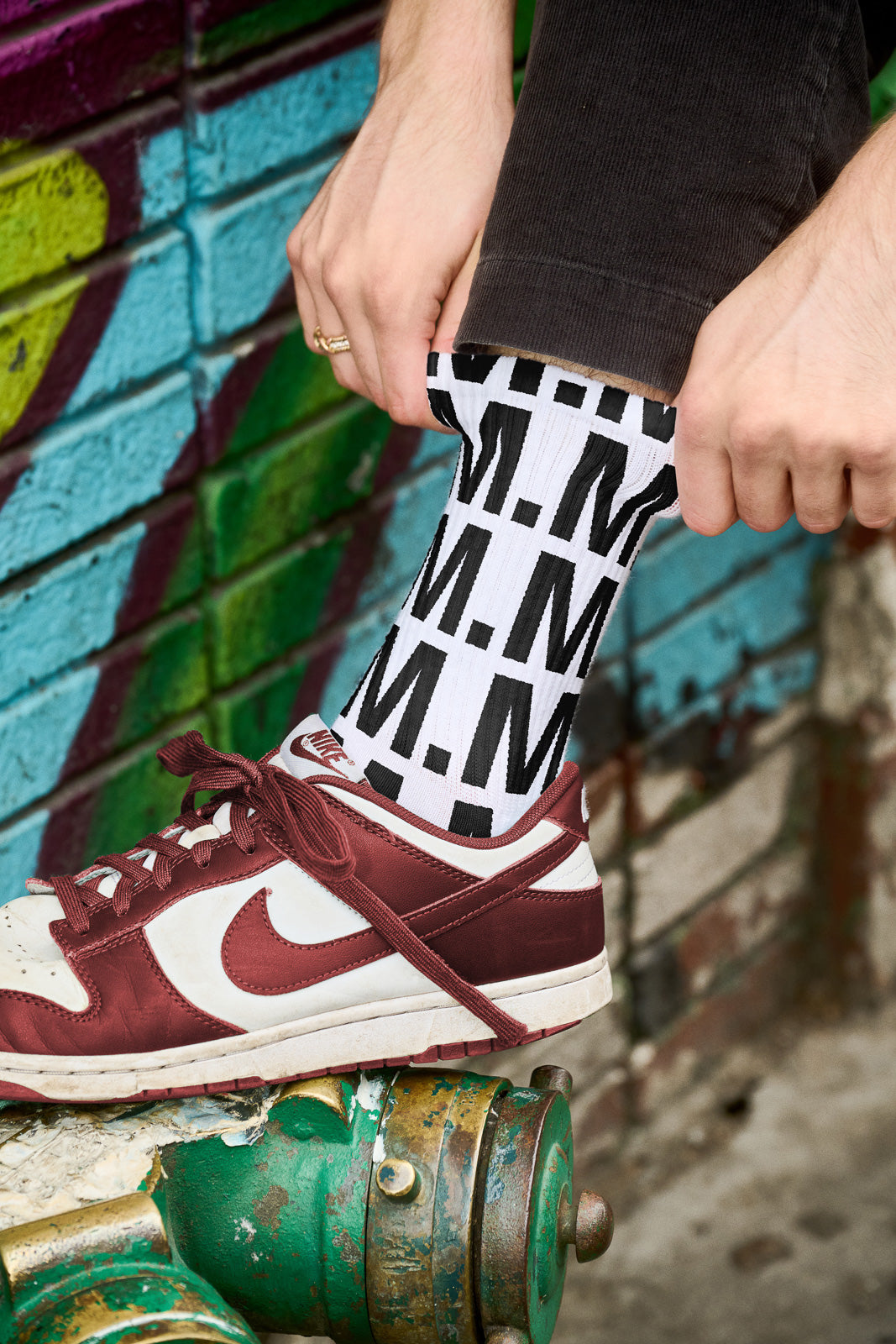 PSD mockup of a person adjusting a white sock with bold, repeating black "M" letters, wearing maroon and white sneakers, against a colorful graffiti wall.