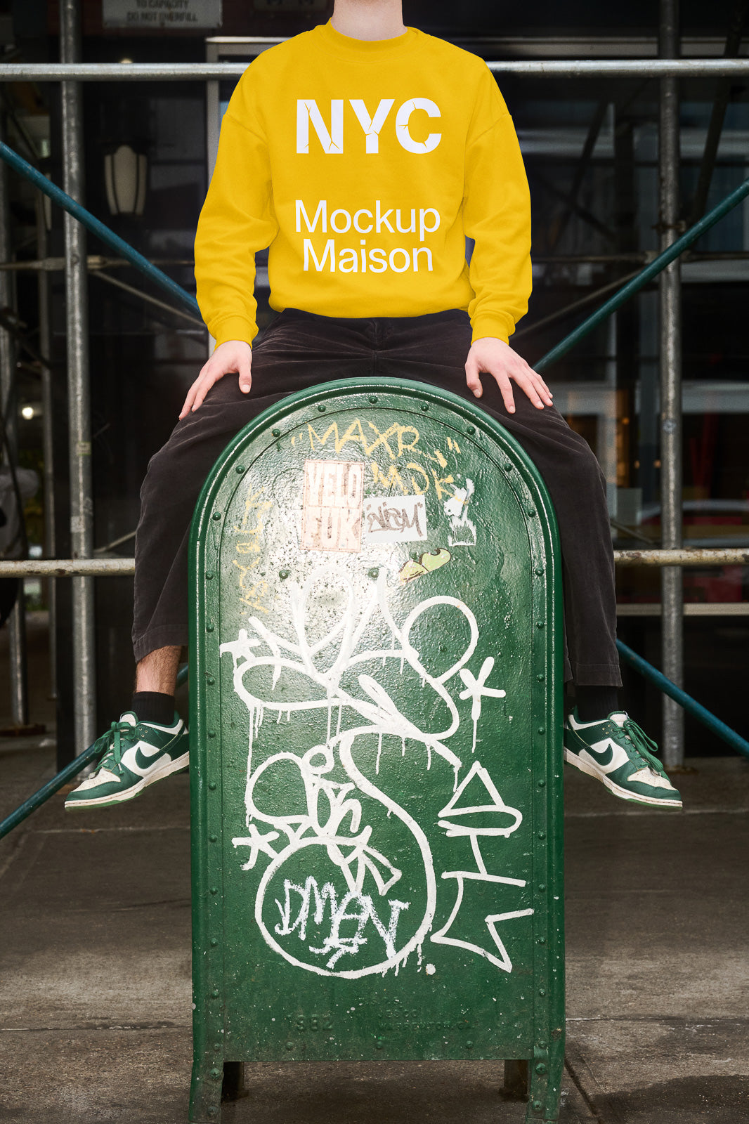 PSD mockup of a person in a yellow sweatshirt labeled "NYC Mockup Maison," sitting on a graffiti-covered green mailbox with visible legs and shoes.