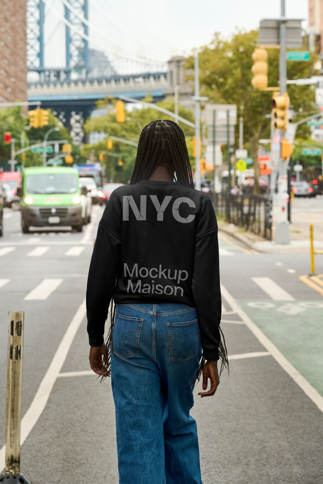 PSD mockup of a person wearing a black sweatshirt with "NYC Mockup Maison" text, walking down an urban street with a bridge in the background.