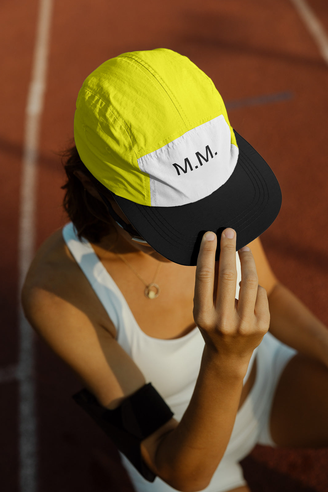 PSD mockup of a person wearing a yellow and white cap with "M.M." printed on it, seated on a track, adjusting the cap's black brim.