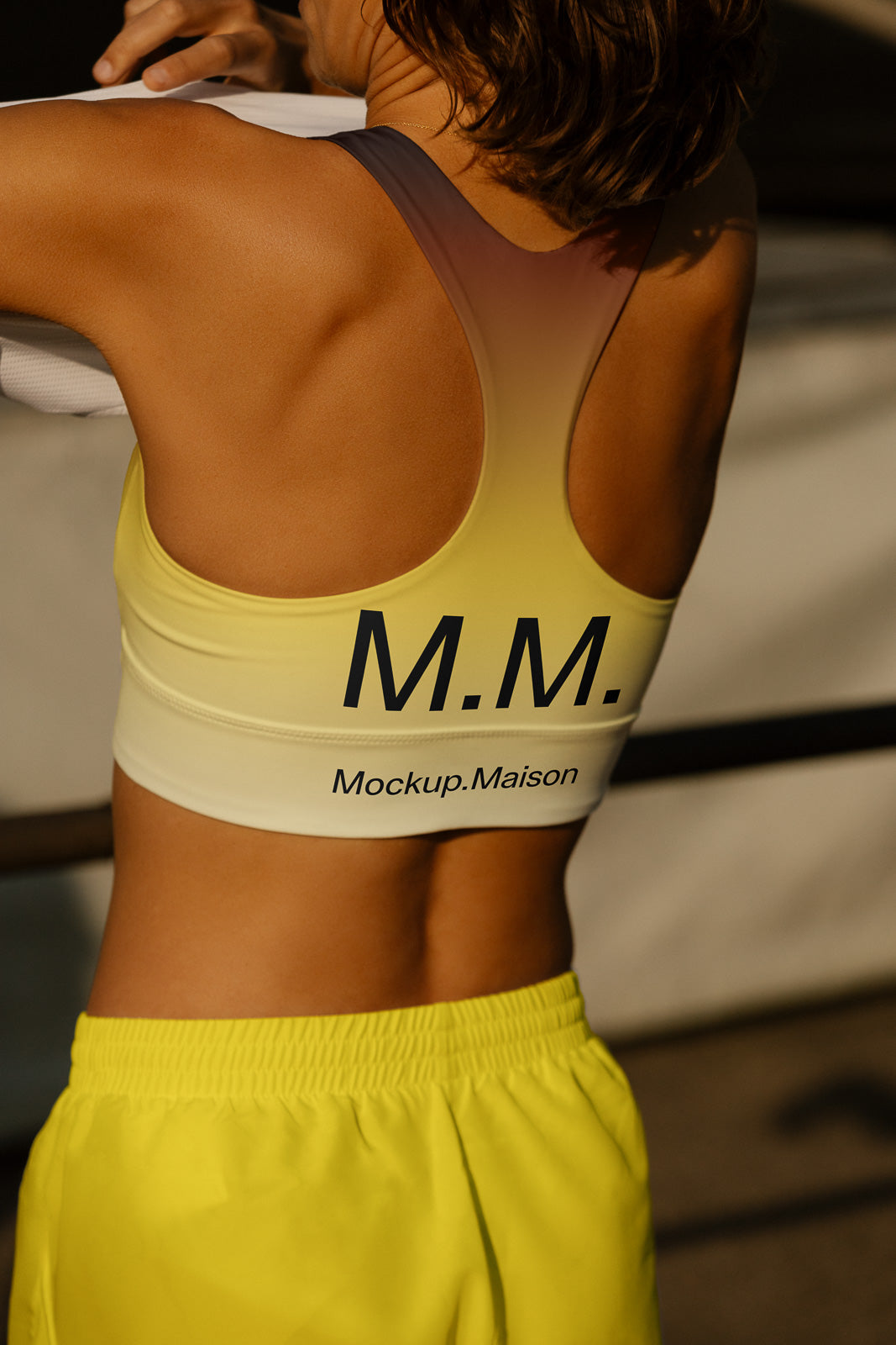 PSD mockup of a person wearing a gradient sports bra with "M.M." and "Mockup.Maison" text on the back, paired with bright yellow shorts.