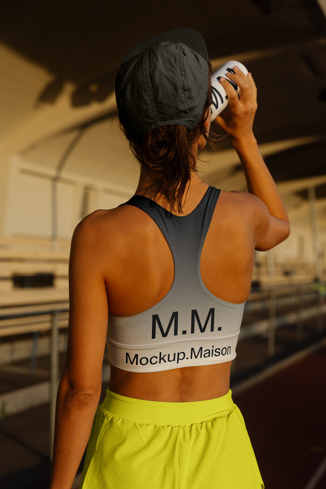 PSD mockup of a person wearing a gray branded sports bra marked "M.M. Mockup.Maison" while drinking from a bottle, standing on a track.