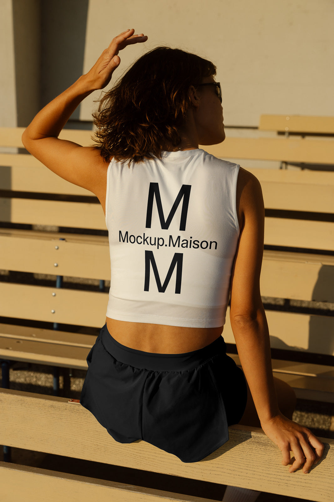 PSD mockup of a person sitting on bleachers wearing a sleeveless top with "Mockup.Maison" and "M" printed on the back, casting a shadow.