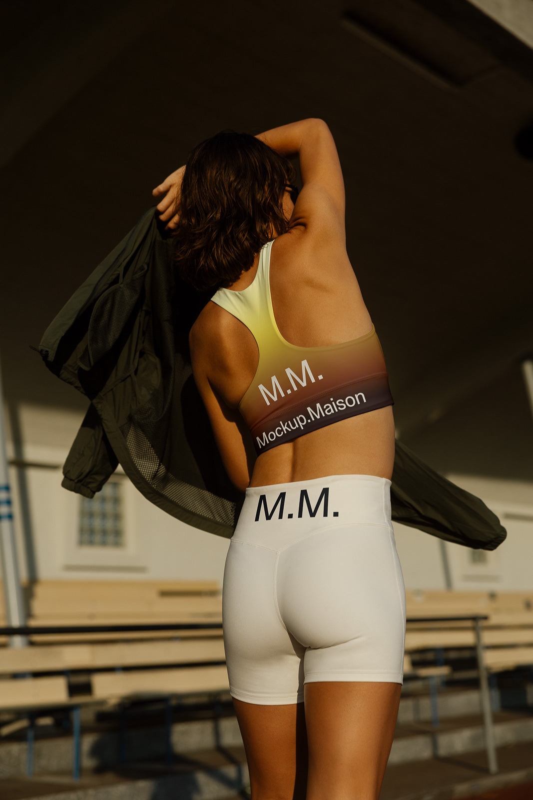 PSD mockup of athletic apparel featuring a woman wearing branded sportswear with "M.M." text, adjusting a jacket. Outdoor setting with benches in the background.
