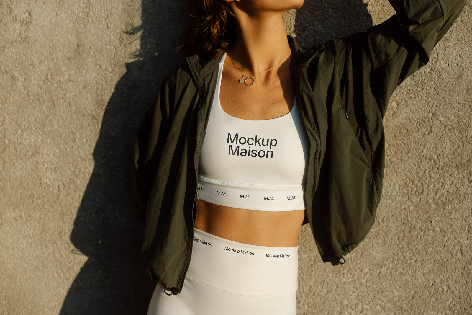 PSD mockup of athletic wear featuring a white sports bra and leggings with "Mockup Maison" logo, worn under a dark jacket against a textured concrete wall.