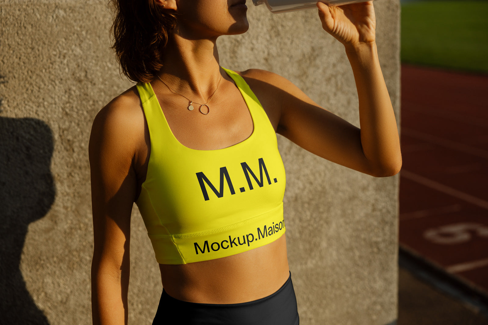 PSD mockup of a woman in a bright yellow sports bra with "M.M." logo, standing outdoors. She's drinking from a bottle, wearing a necklace with round charms.