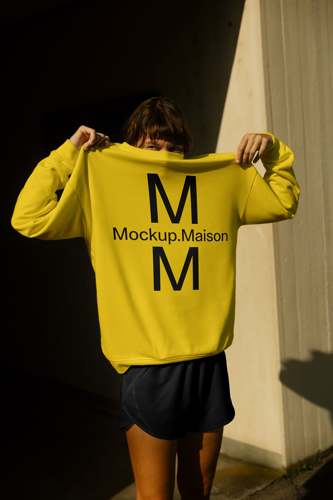 PSD mockup of a yellow sweatshirt with "Mockup.Maison" text, held up in front of a person's face, contrasted against a shadowed background.