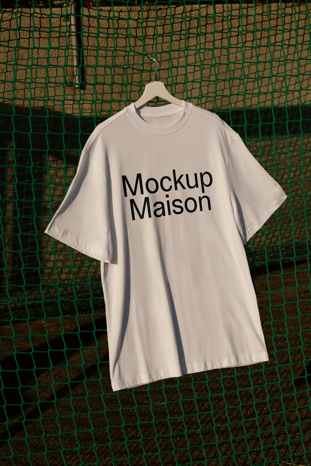 PSD mockup of a white T-shirt with "Mockup Maison" text, hanging on a hanger against a green net backdrop under natural light.
