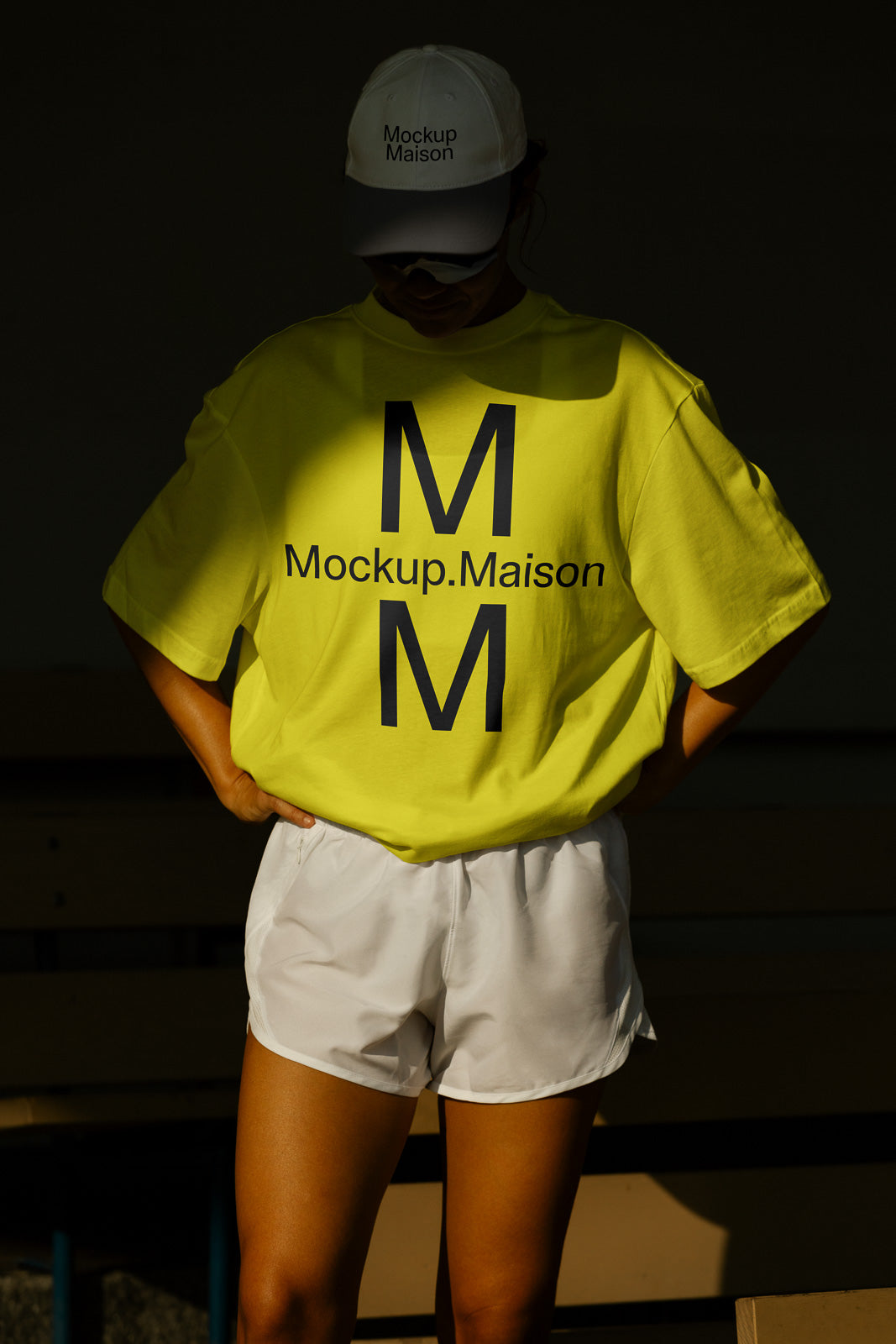 PSD mockup of a person wearing a bright yellow shirt and white shorts, with "Mockup.Maison" text and logo in a shadowed setting.