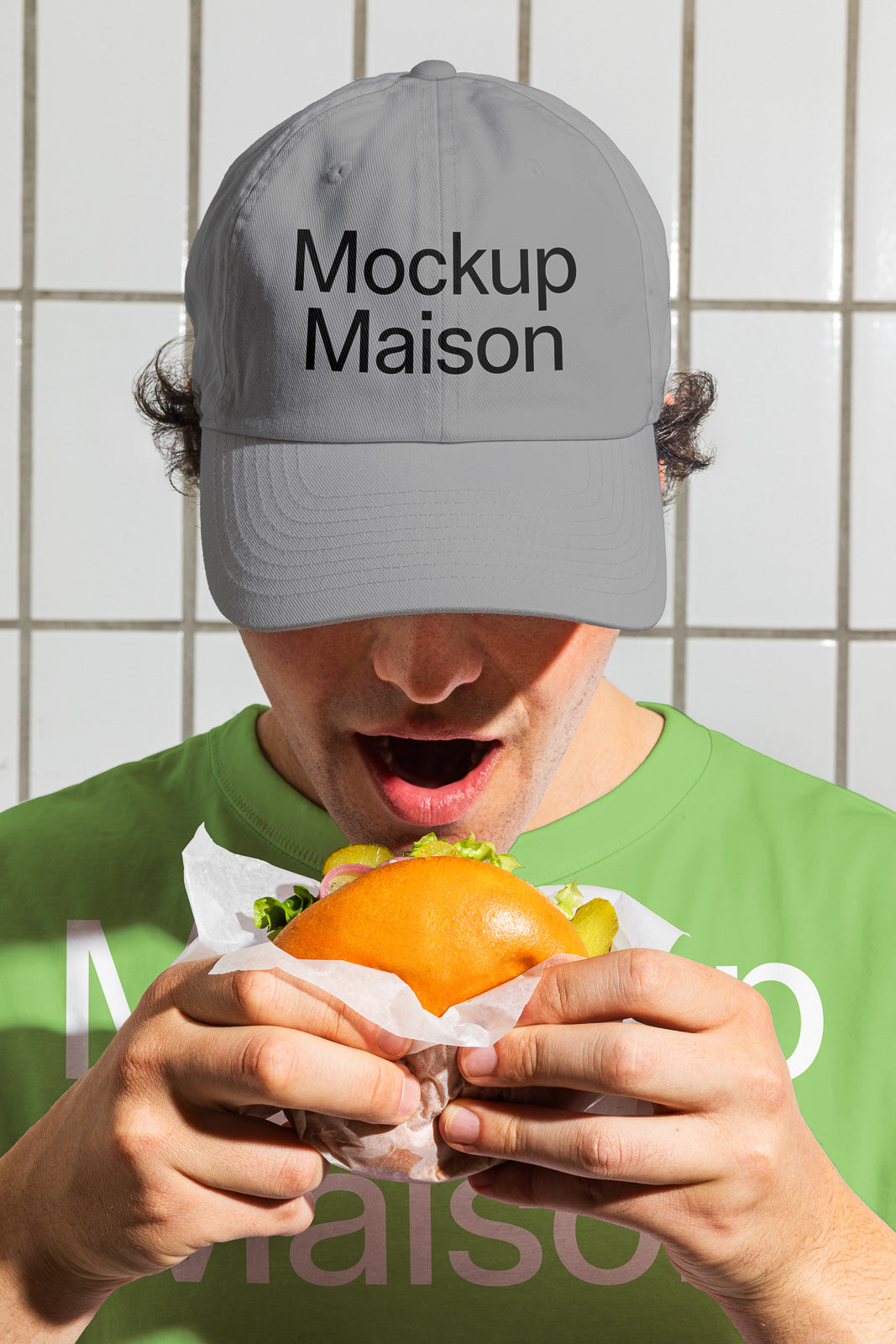 PSD mockup of a person wearing a gray cap with "Mockup Maison" text, holding a burger wrapped in paper, wearing a green shirt in front of a tiled wall.