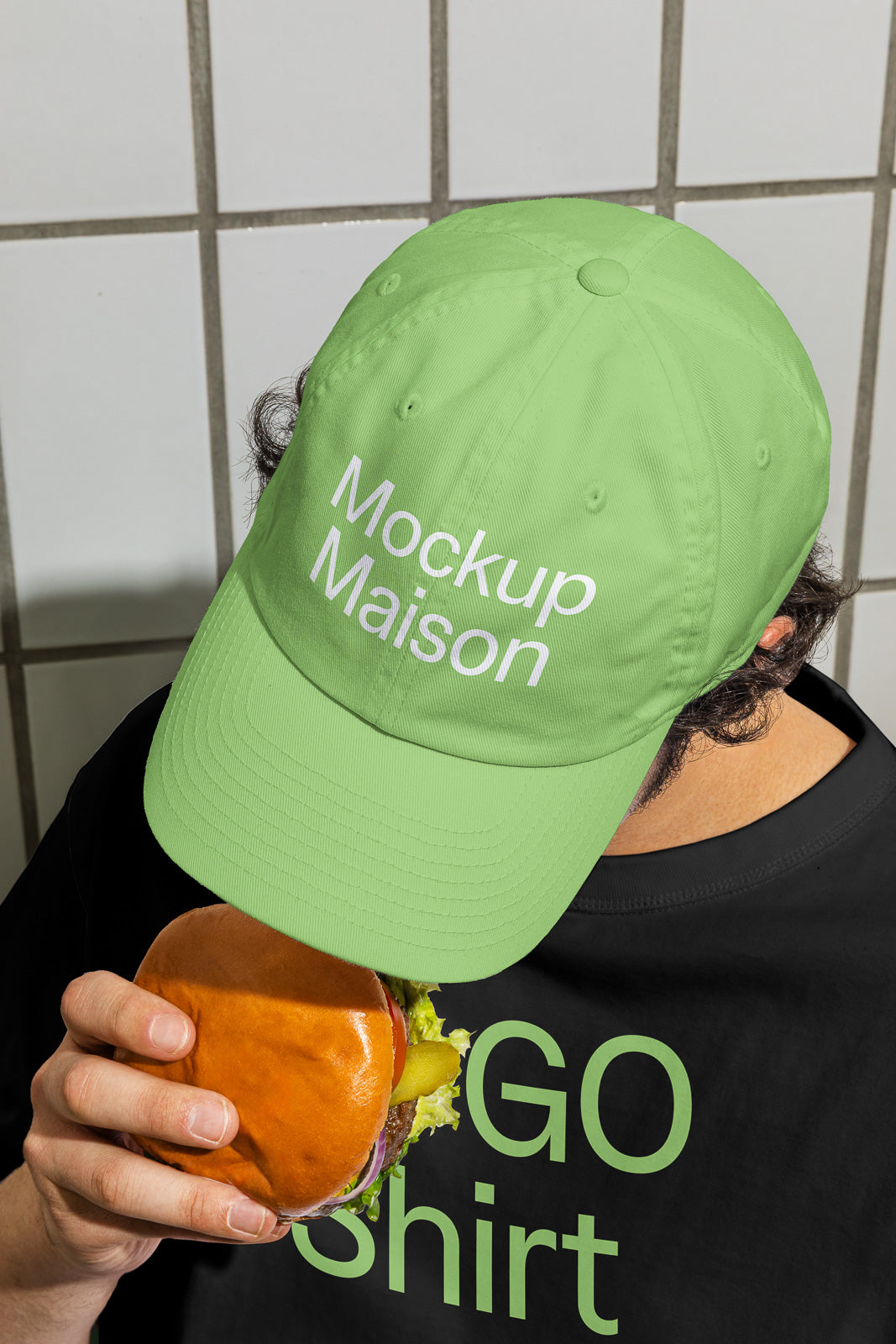 PSD mockup of a person in a green cap with "Mockup Maison" text, holding a burger in one hand, wearing a black shirt with green lettering.