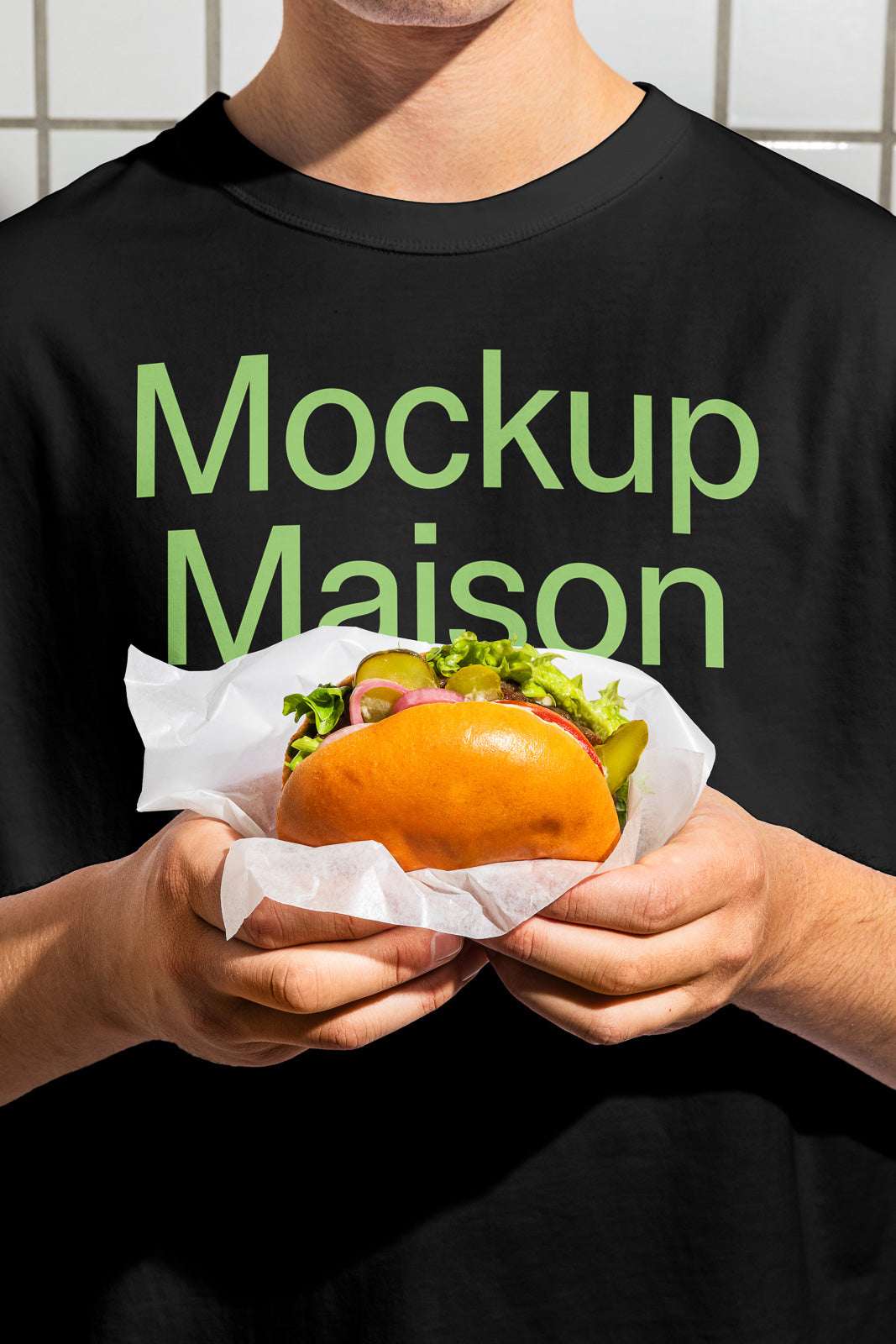 PSD mockup of a person holding a burger wrapped in paper, wearing a black shirt with green text saying "Mockup Maison."