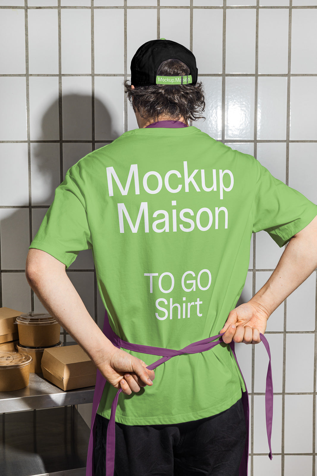 PSD mockup of a person wearing a green "Mockup Maison TO GO Shirt" with a purple apron, standing in a tiled kitchen setting with takeaway boxes.