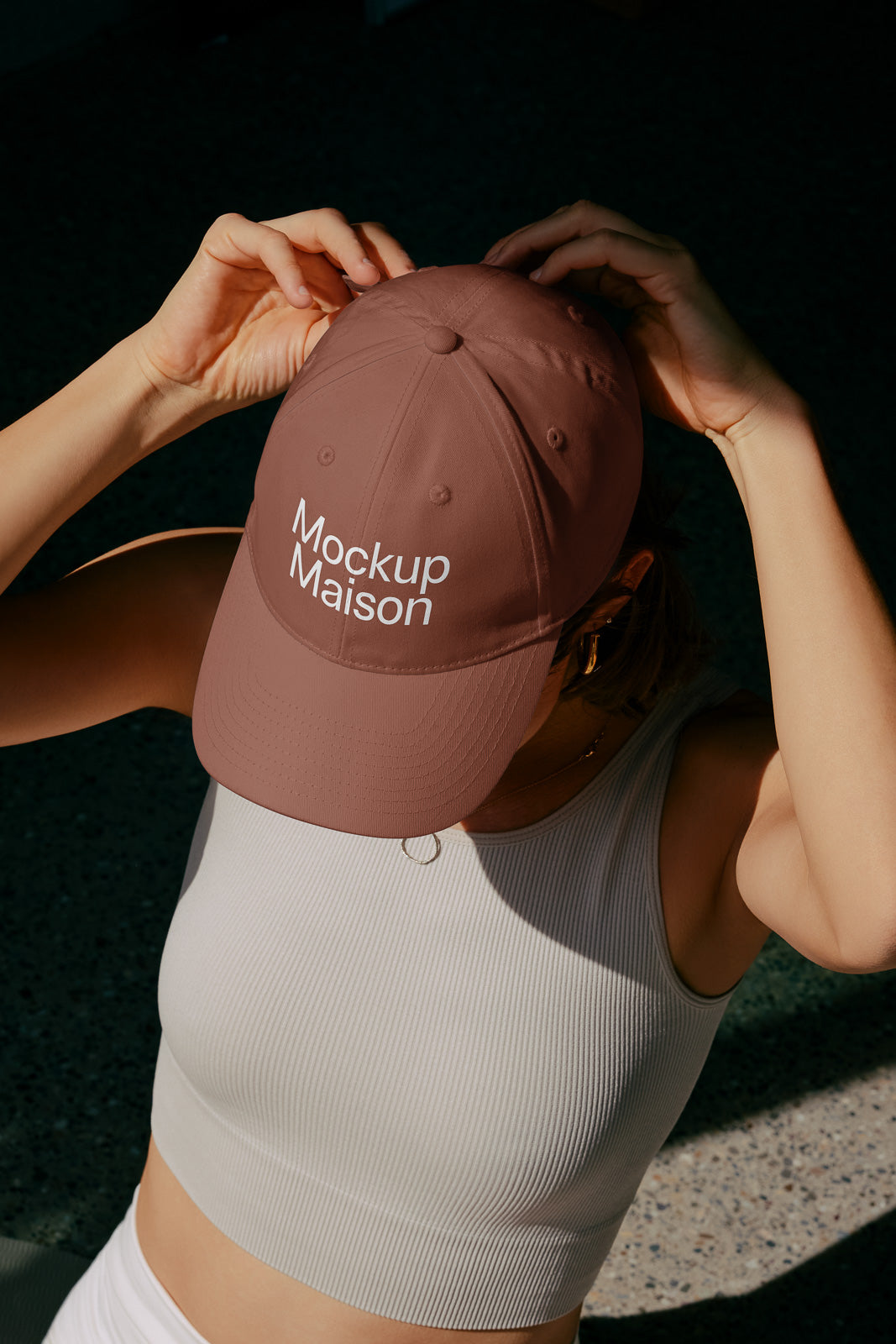 PSD mockup of a person adjusting a brown cap with "Mockup Maison" text, wearing a light sleeveless top against a shaded background.
