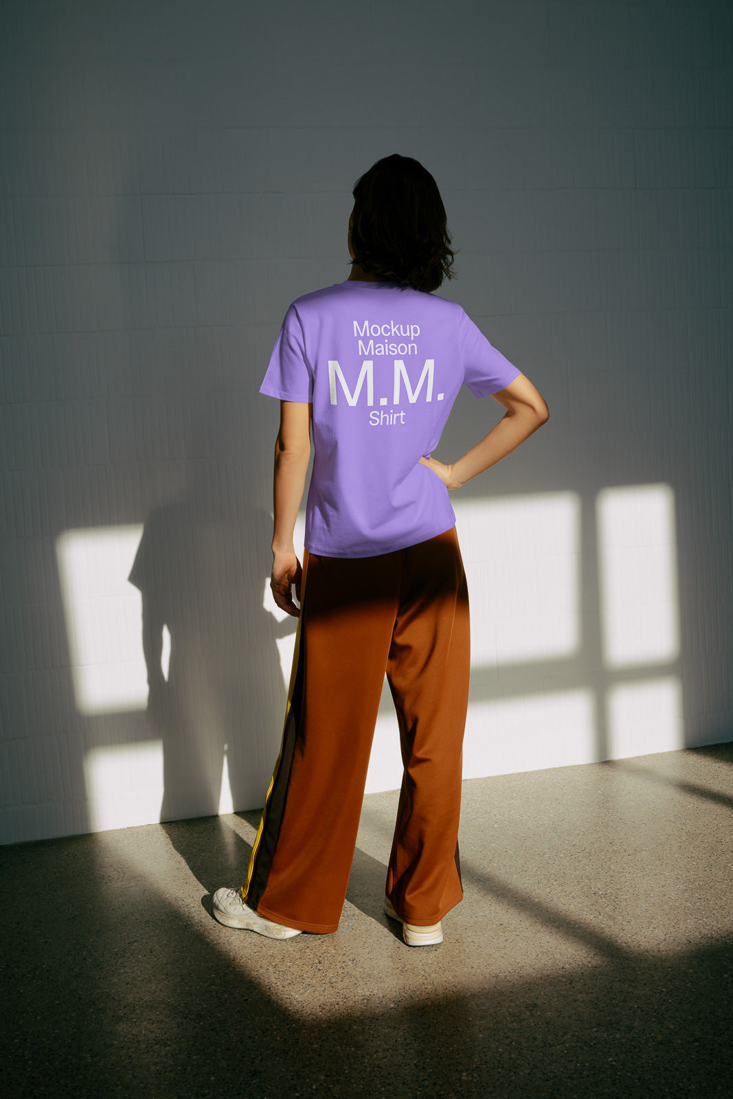 PSD mockup of a person wearing a purple shirt with "Mockup Maison M.M. Shirt" text, posing with hand on hip against a sunlit wall.
