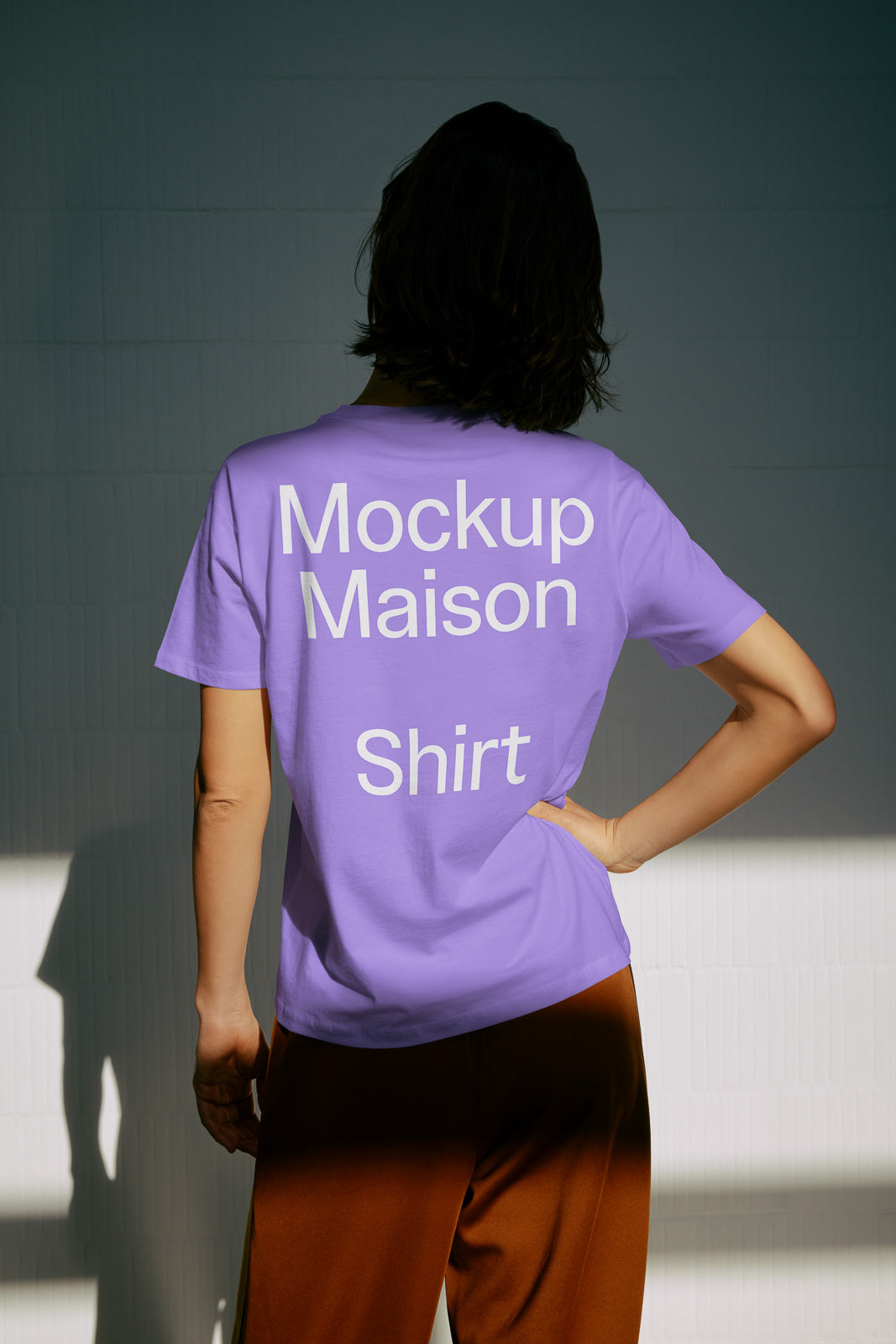 PSD mockup of a person wearing a purple T-shirt with "Mockup Maison Shirt" text on the back, standing indoors against a textured wall.
