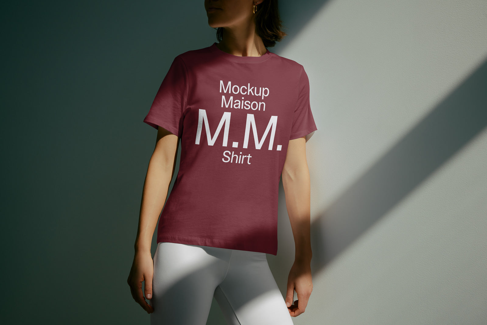 PSD mockup of a person wearing a maroon t-shirt labeled with "Mockup Maison M.M. Shirt" in a well-lit setting, casting distinct shadows on the wall.