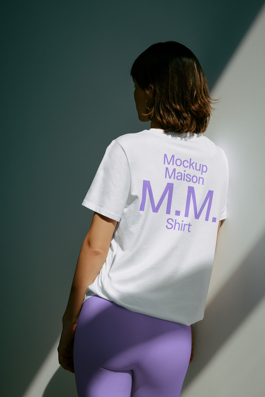 PSD mockup of a woman wearing a white t-shirt with purple text "Mockup Maison M.M. Shirt," paired with purple leggings, standing against a wall.