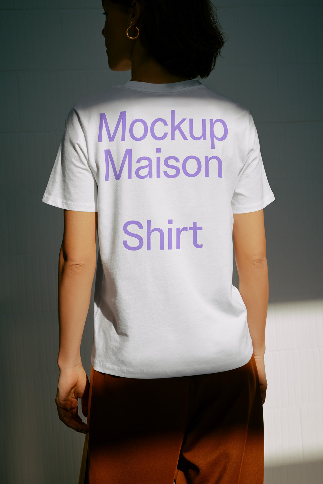 PSD mockup of a white t-shirt worn by a person, displaying purple text "Mockup Maison Shirt" on the back, set against a shadowed backdrop.