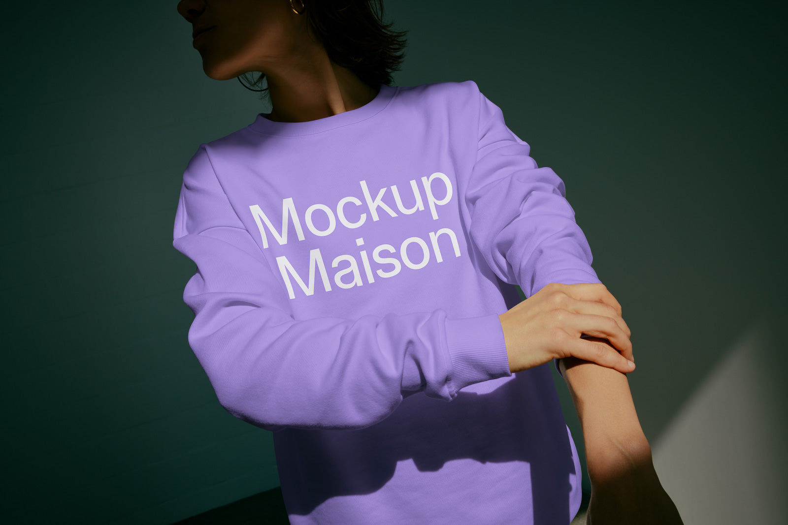 PSD mockup of a person wearing a purple sweatshirt with "Mockup Maison" text, folding their sleeve in a dimly lit setting.