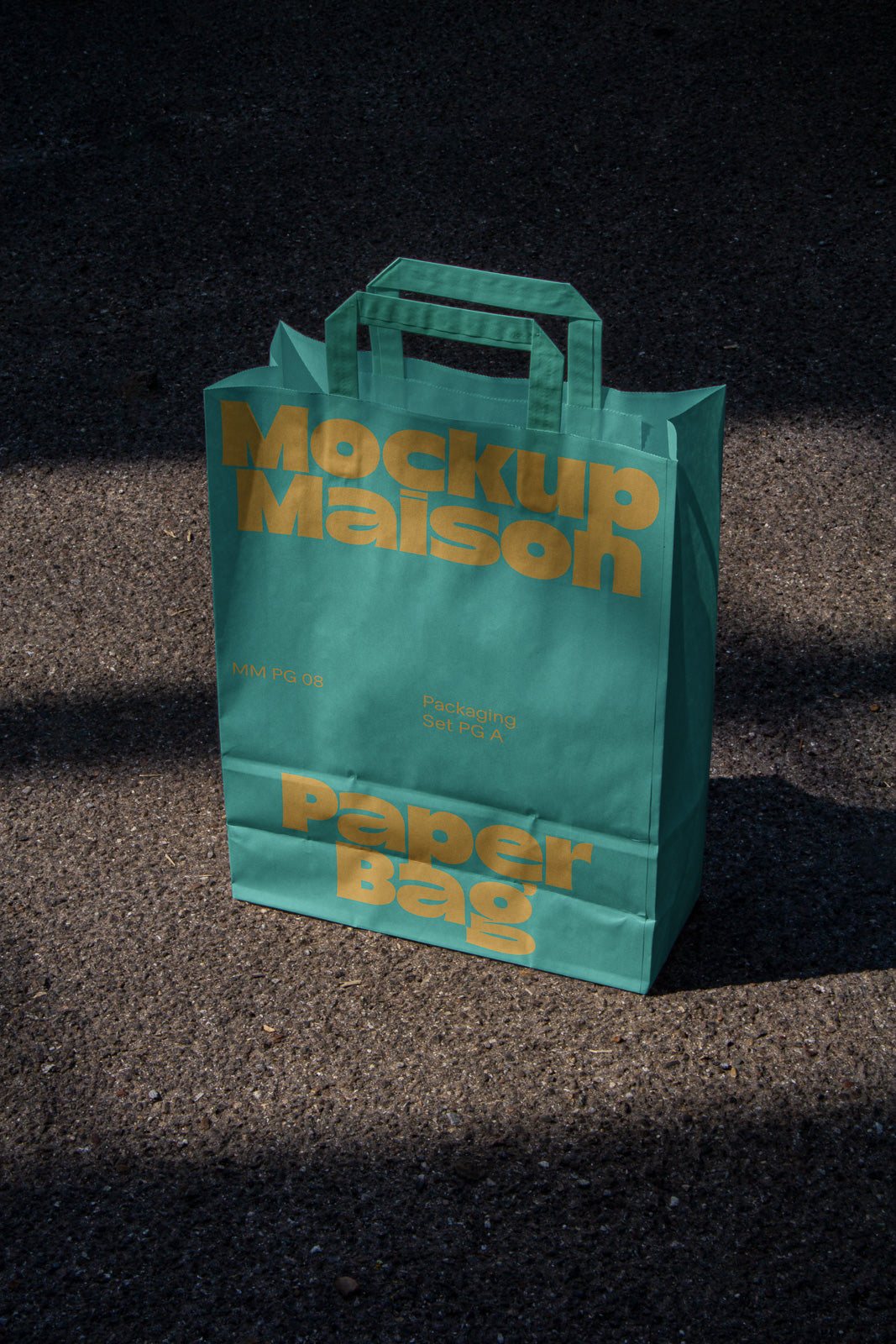 PSD mockup of a teal paper bag with gold lettering that reads "Mockup Maison Paper Bag," displayed on a textured asphalt surface.
