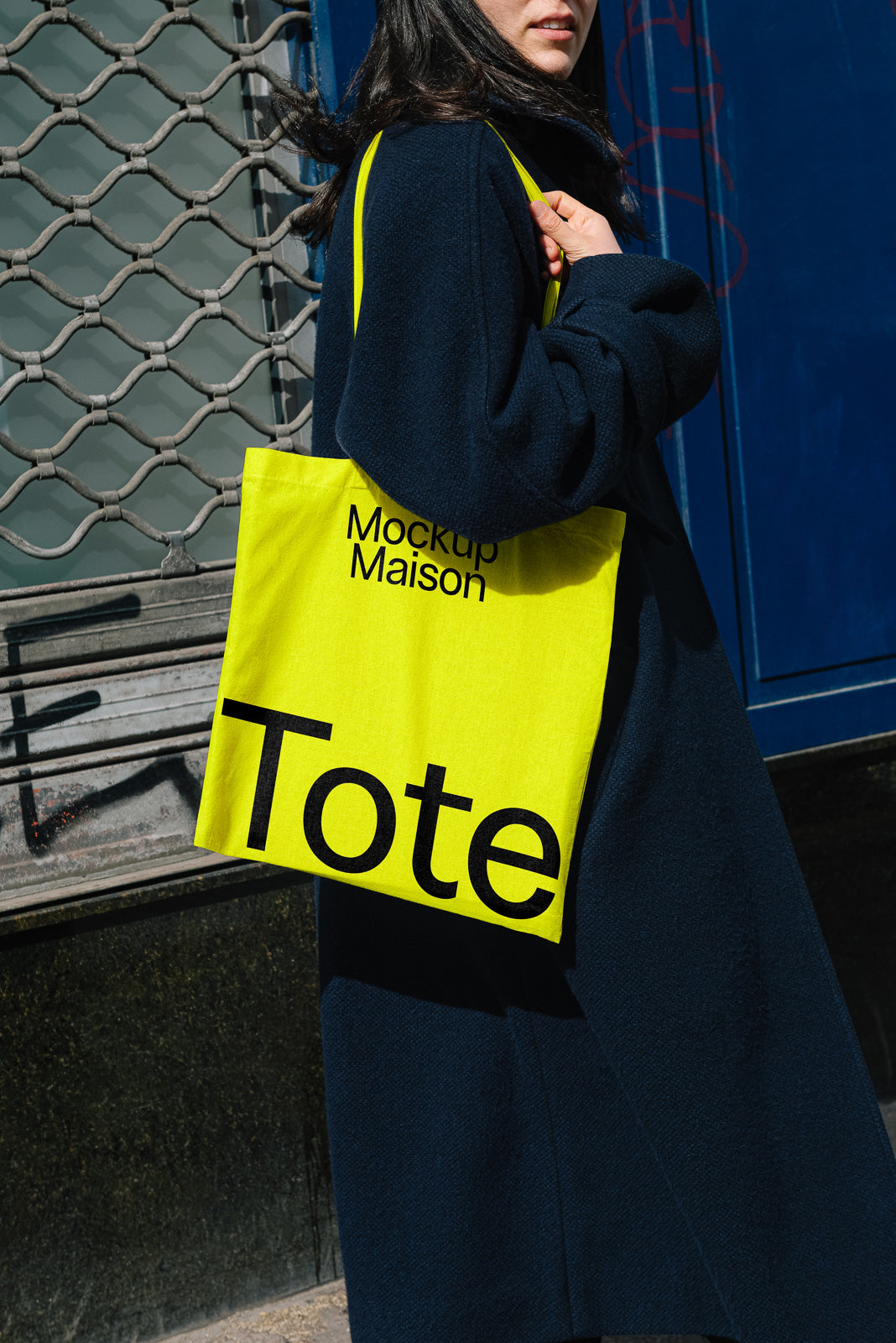 PSD mockup of a bright yellow tote bag with "Mockup Maison" and "Tote" text, held by a person in dark clothing, against a blue door and metal gate.