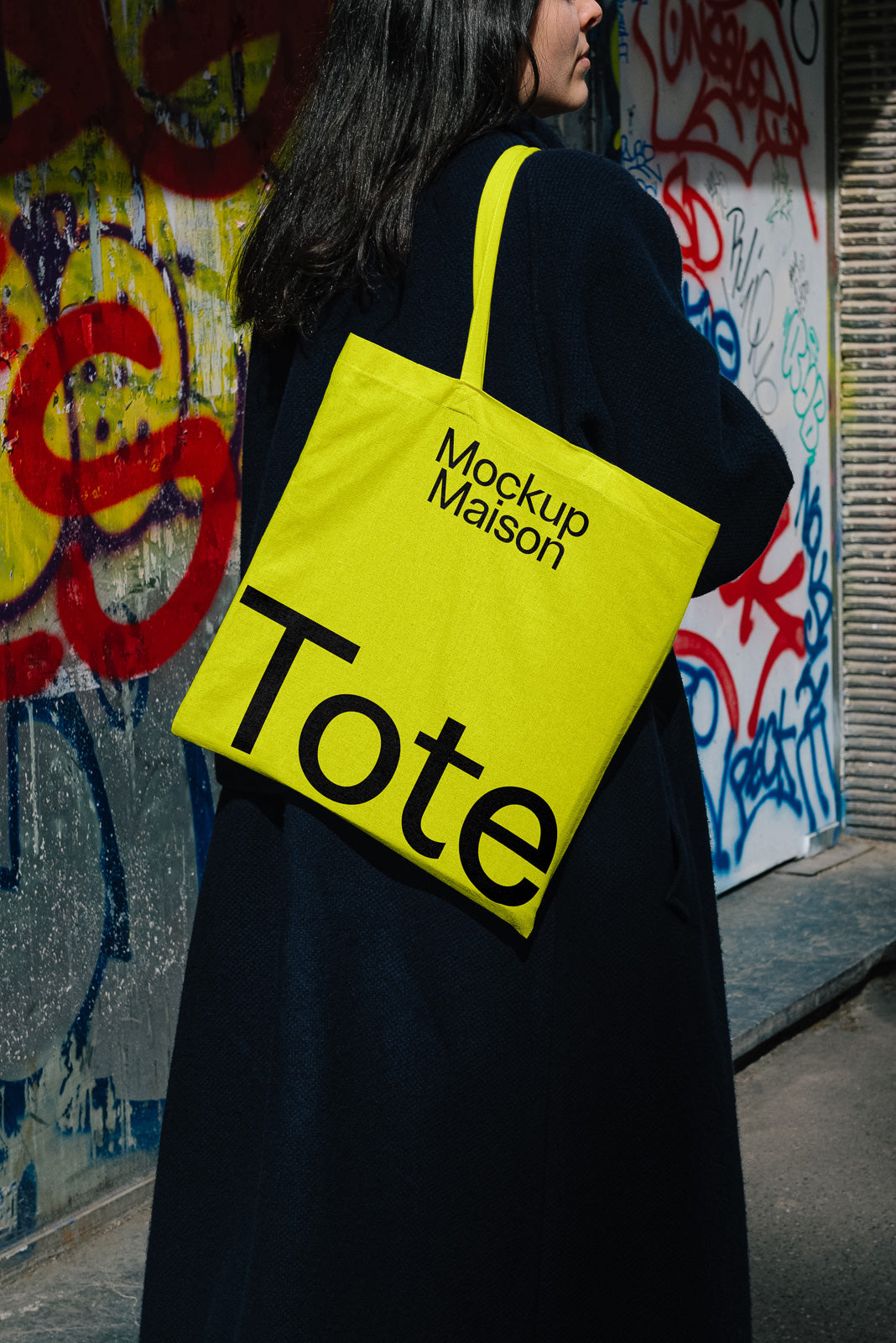 PSD mockup of a woman carrying a bright yellow tote bag with "Mockup Maison Tote" printed on it, standing against a graffiti-covered wall.