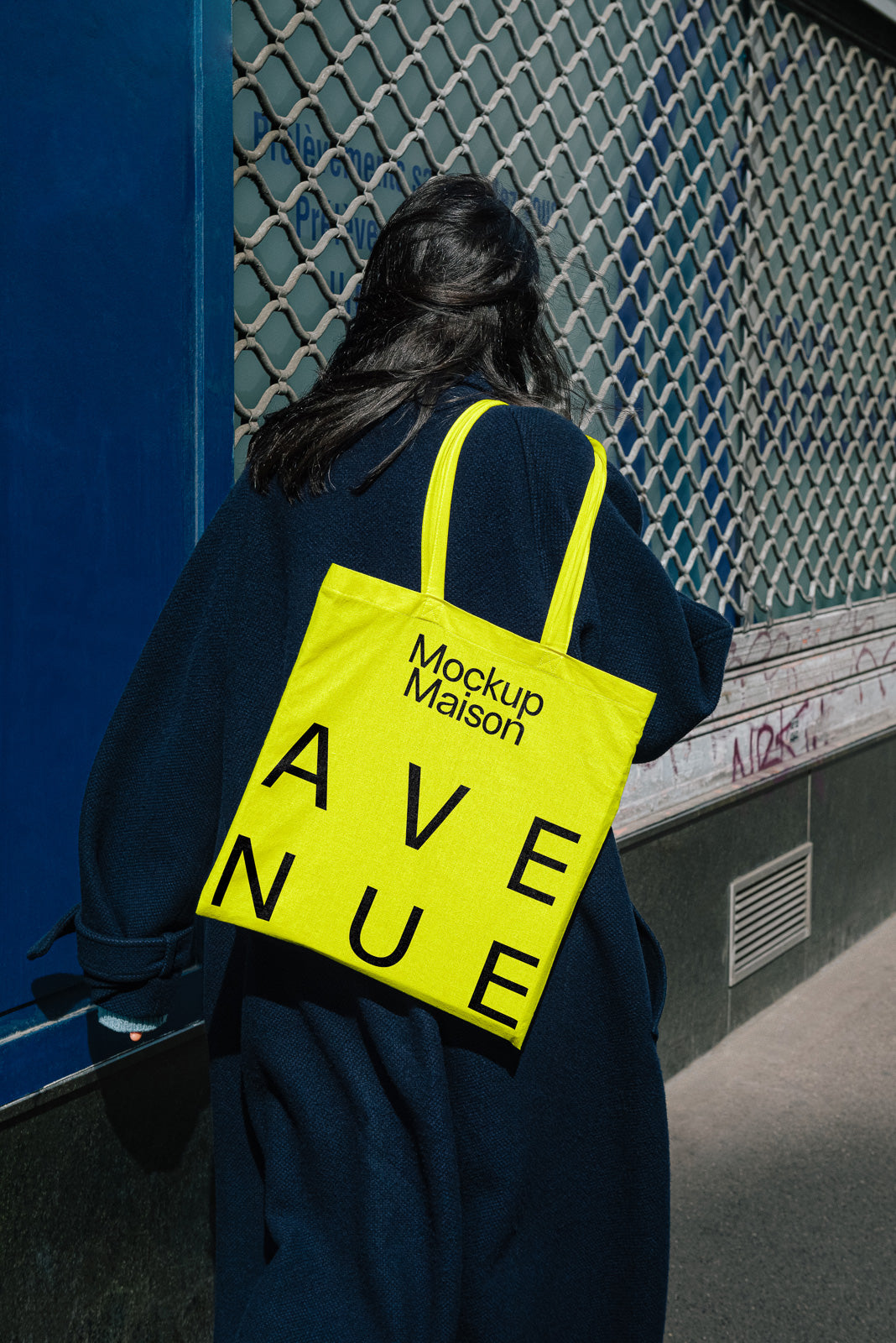 PSD mockup of a person carrying a yellow tote bag with bold black text "Mockup Maison AVENUE," standing near a blue wall with a metal grate.