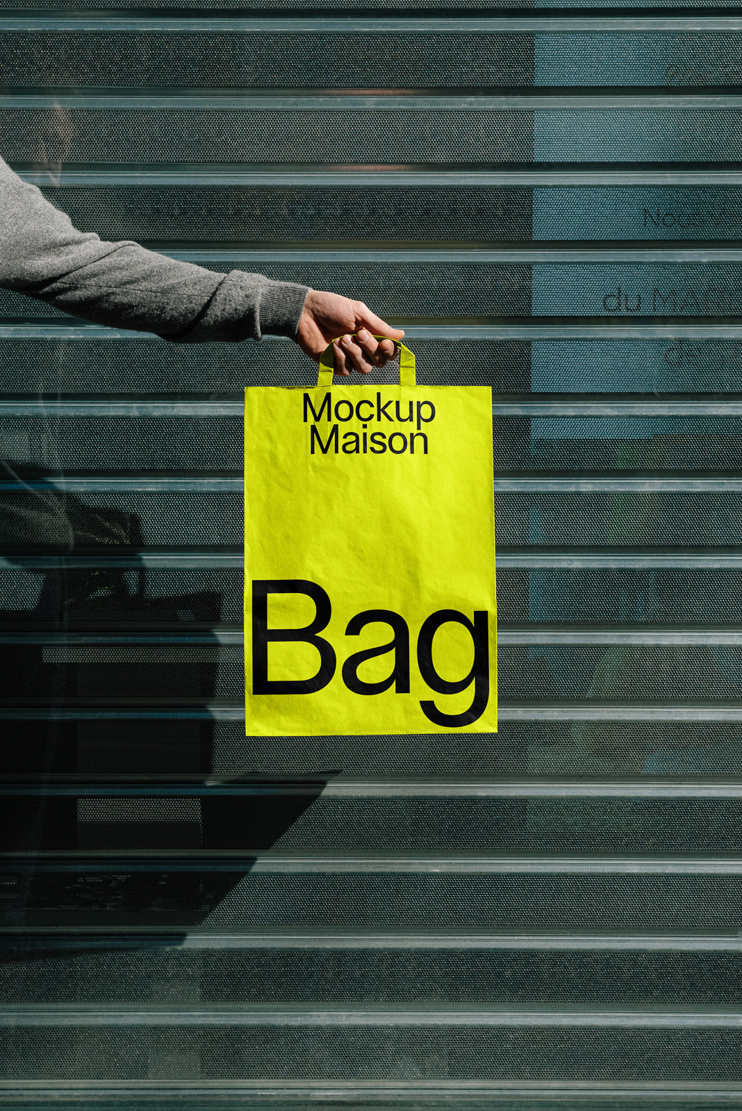 PSD mockup of a hand holding a bright yellow paper bag labeled "Mockup Maison Bag" against a gray striped background.