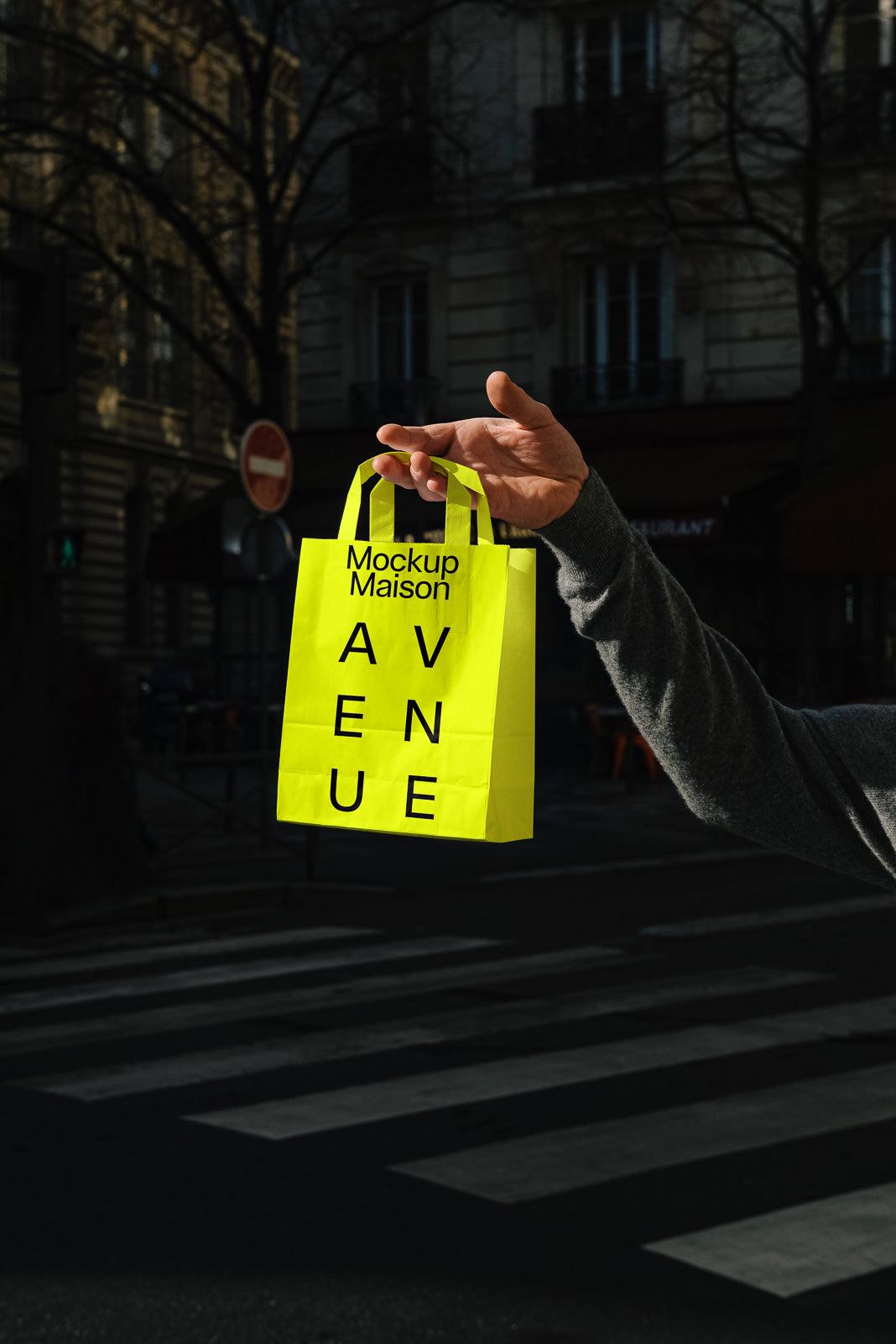 PSD mockup of a hand holding a bright yellow shopping bag with "Mockup Maison AVENUE" printed in black, against a city street background.