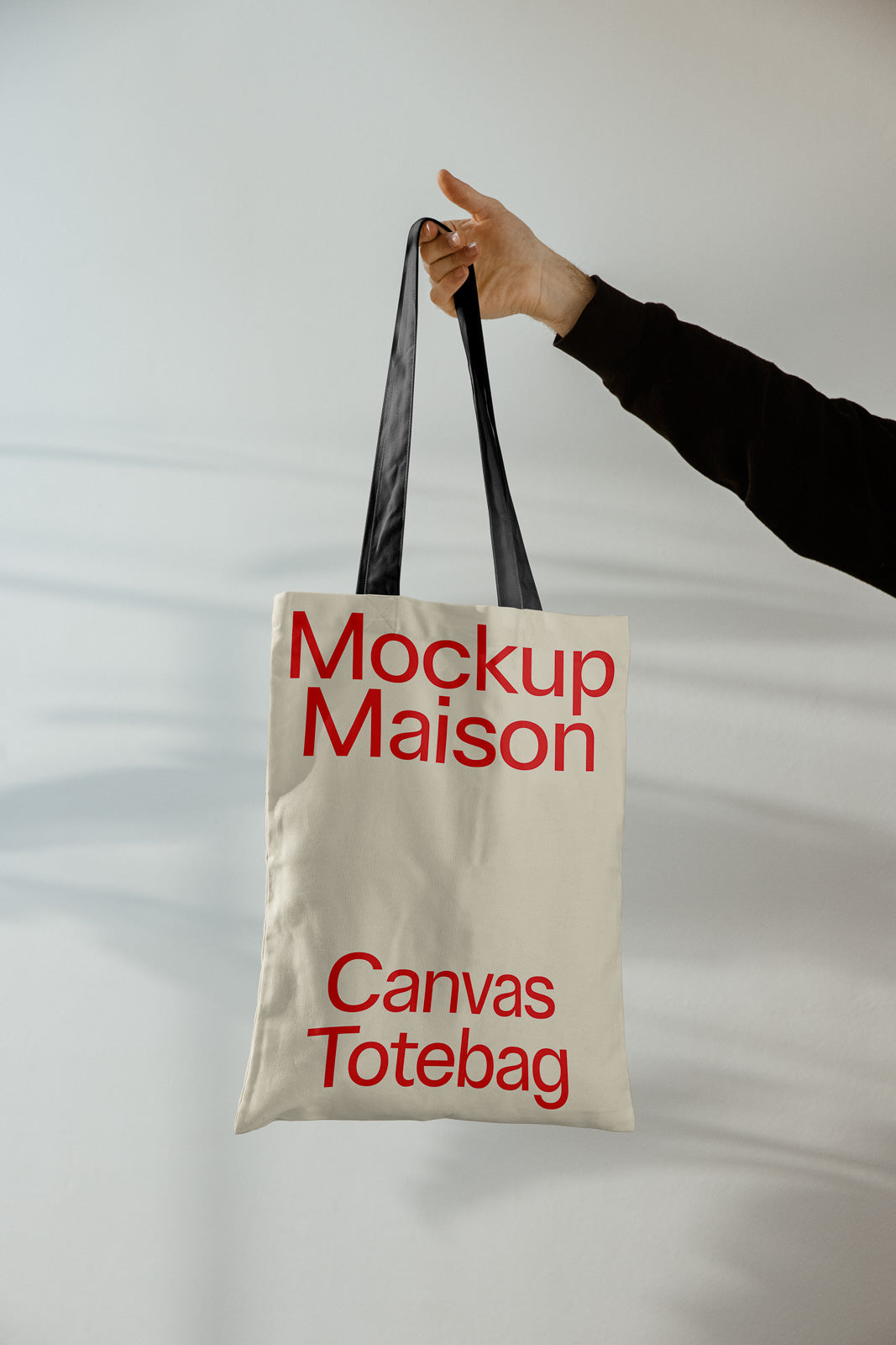 PSD mockup of a canvas tote bag being held by a hand, displaying bold red text "Mockup Maison" and "Canvas Totebag" on a neutral background.