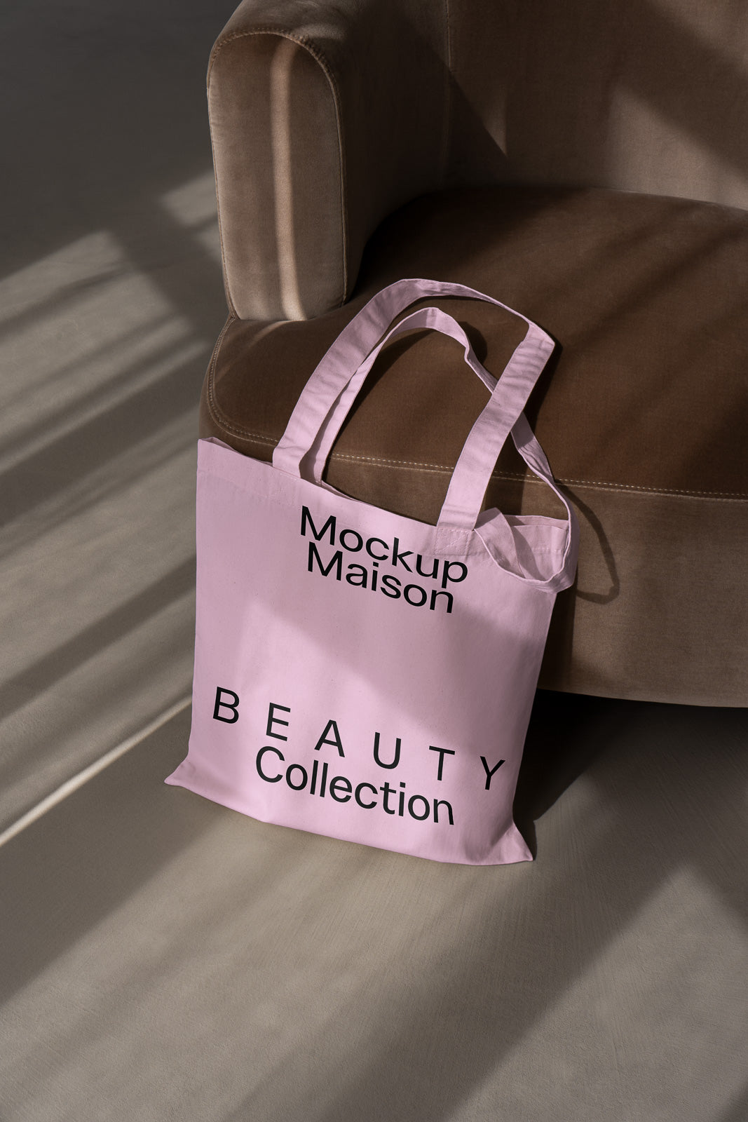 PSD mockup of a pink tote bag titled "Mockup Maison Beauty Collection" placed on a brown sofa with light and shadows creating a textured backdrop.