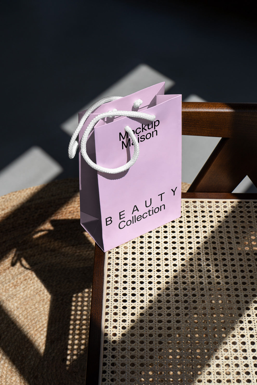 PSD mockup of a lavender paper shopping bag with the text "Mockup Maison" and "Beauty Collection," placed on a woven chair with sunlight casting shadows.
