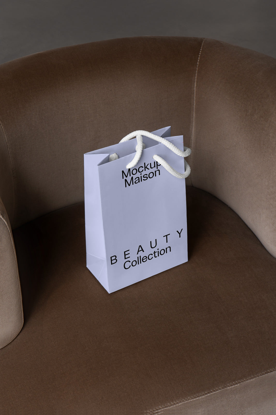 PSD mockup of a white paper shopping bag with "Mockup Maison" and "Beauty Collection" text, placed on a brown upholstered chair.