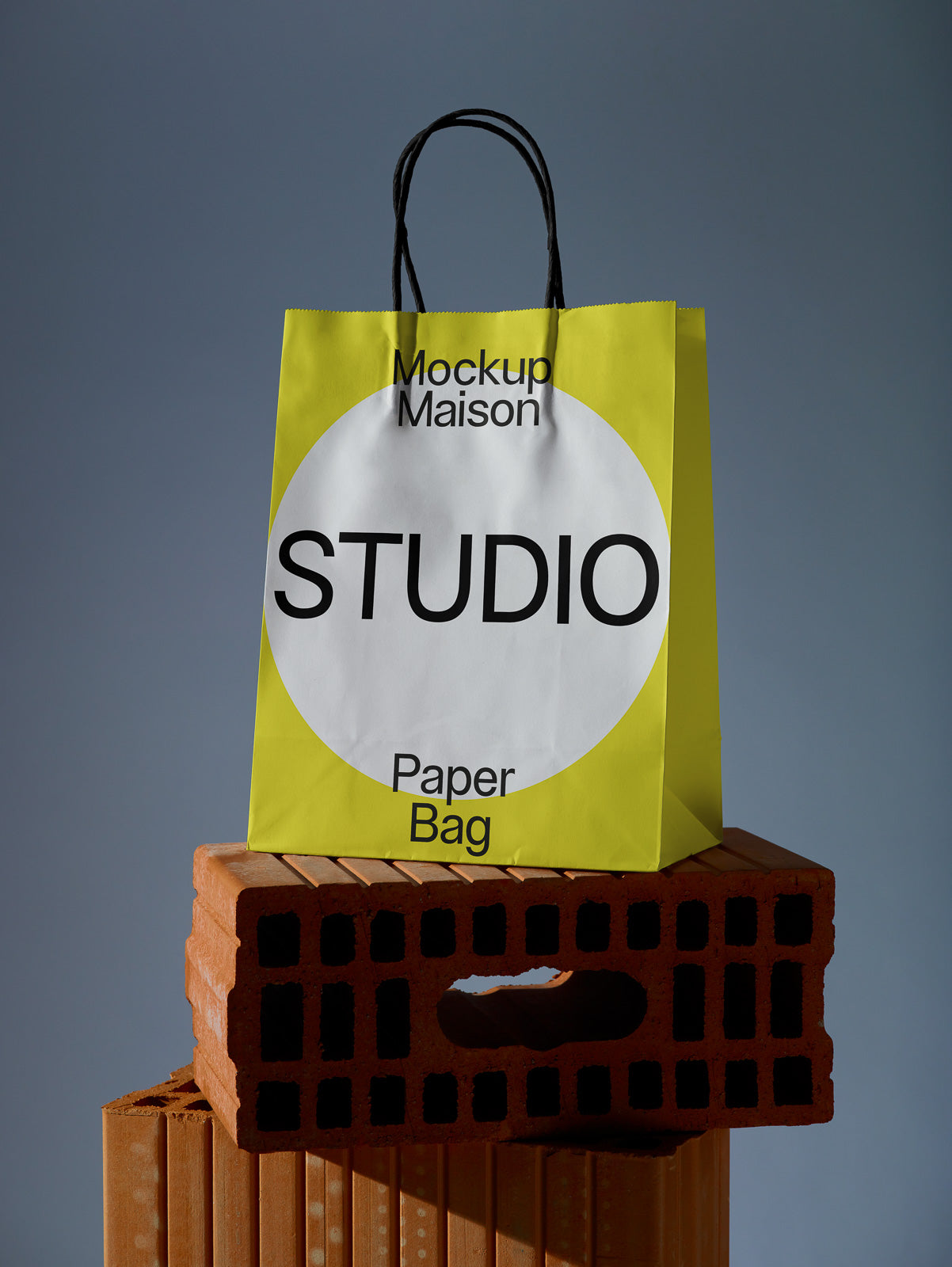 PSD mockup of a yellow paper bag with black handles and large circular white label, placed on stacked terracotta bricks against a gray background.