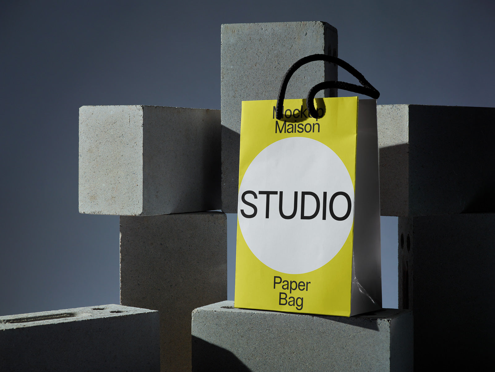 PSD mockup of a yellow paper bag with a white circle, black text "STUDIO," placed against gray concrete blocks.