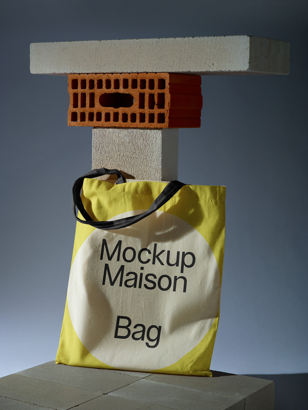 PSD mockup of a yellow tote bag with "Mockup Maison Bag" text, leaning against concrete and brick slabs, creating an industrial aesthetic.