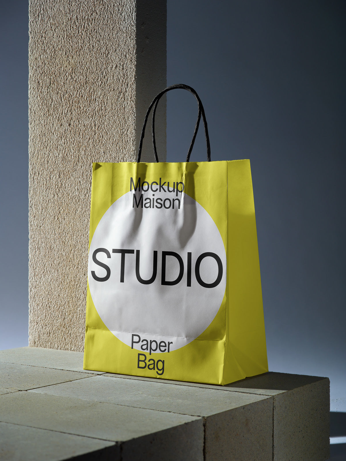PSD mockup of a yellow paper bag with black handles, featuring the words “Mockup Maison” and “STUDIO” in bold lettering, placed on a textured block.